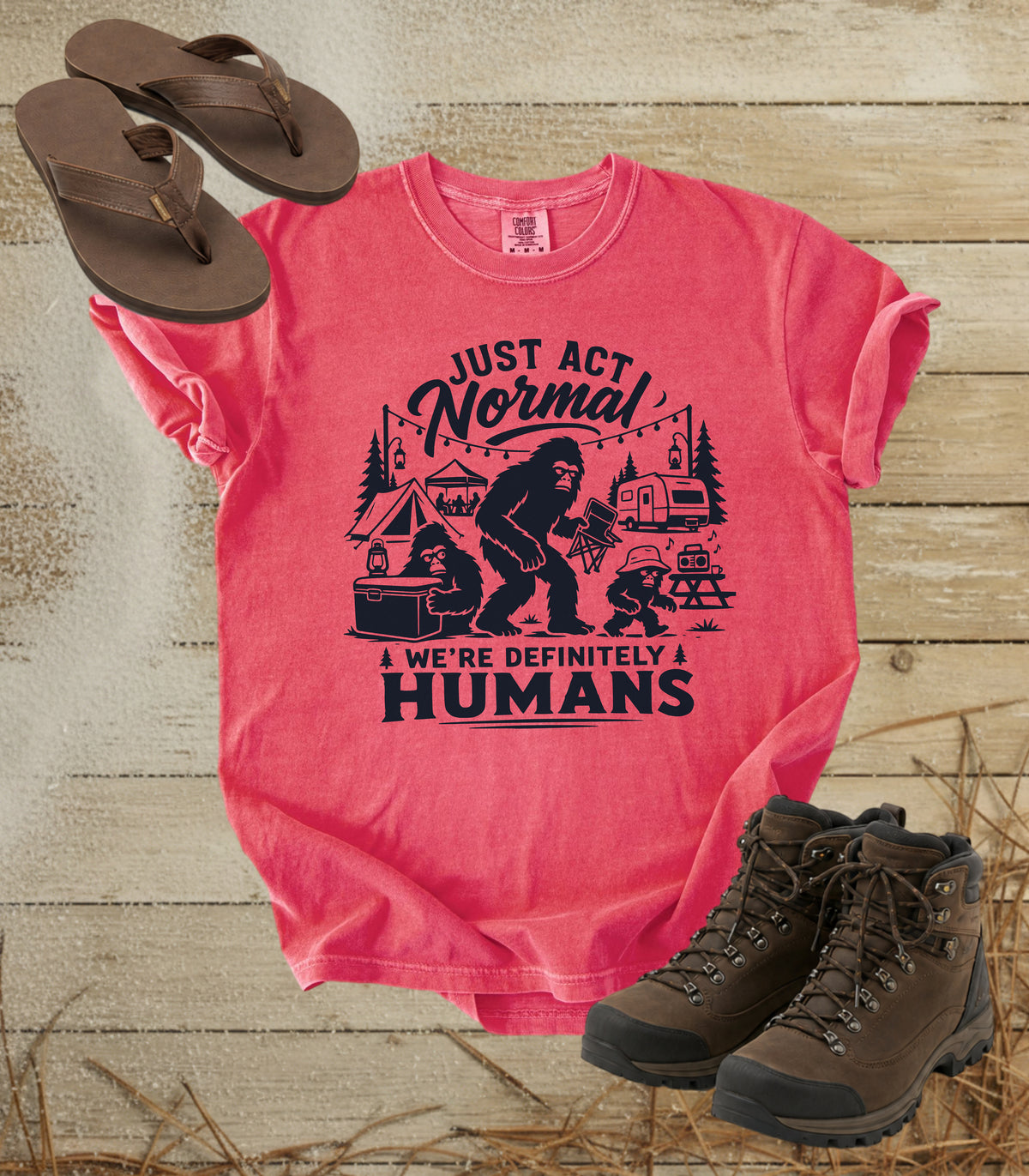 A comfort colors watermelon color graphic t-shirt featuring a dark print of a Bigfoot family camping scene with the text 'JUST ACT Normal WE'RE DEFINITELY HUMANS'. The shirt is styled in a flat-lay arrangement on a rustic wooden background, accompanied by brown leather sandals and hiking boots.