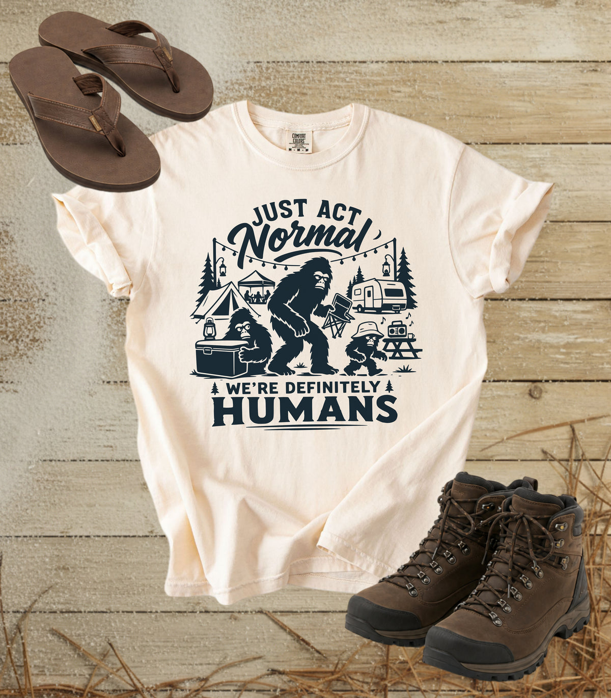 A comfort colors ivory color graphic t-shirt featuring a dark print of a Bigfoot family camping scene with the text 'JUST ACT Normal WE'RE DEFINITELY HUMANS'. The shirt is styled in a flat-lay arrangement on a rustic wooden background, accompanied by brown leather sandals and hiking boots.