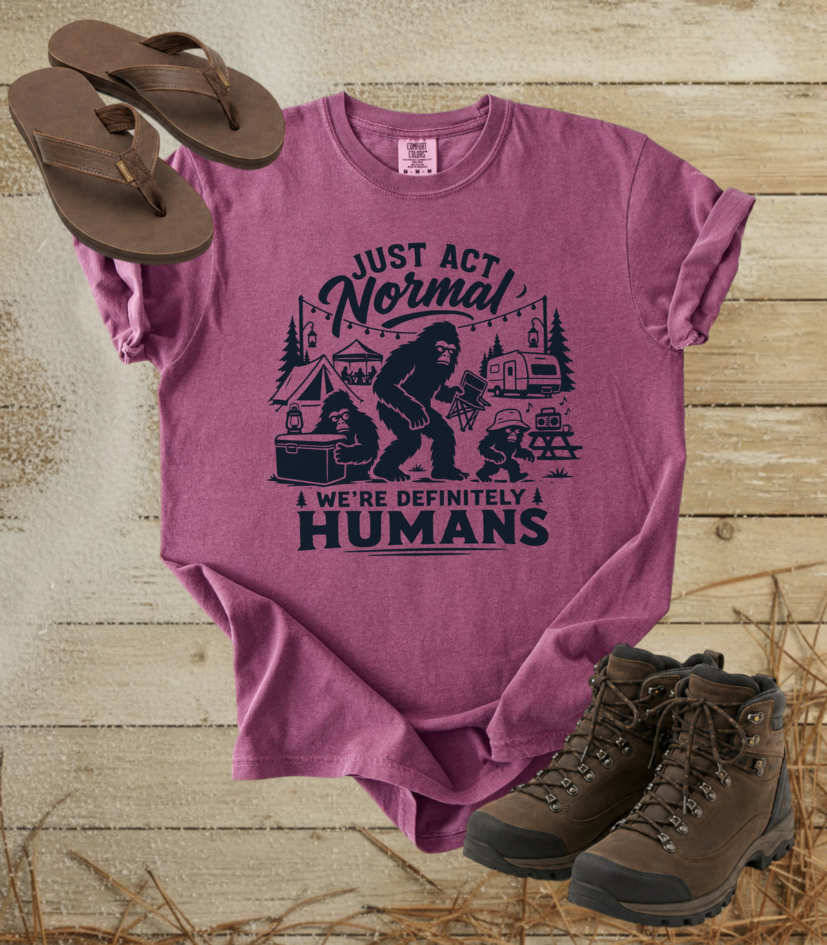 A comfort colors berry color graphic t-shirt featuring a dark print of a Bigfoot family camping scene with the text 'JUST ACT Normal WE'RE DEFINITELY HUMANS'. The shirt is styled in a flat-lay arrangement on a rustic wooden background, accompanied by brown leather sandals and hiking boots.