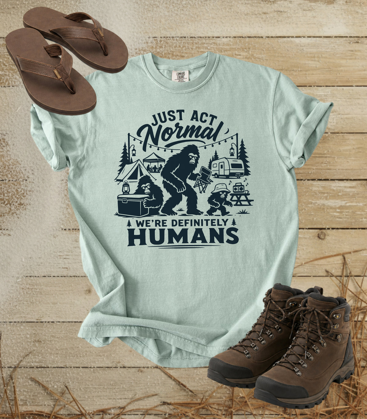 A comfort colors bay color graphic t-shirt featuring a dark print of a Bigfoot family camping scene with the text 'JUST ACT Normal WE'RE DEFINITELY HUMANS'. The shirt is styled in a flat-lay arrangement on a rustic wooden background, accompanied by brown leather sandals and hiking boots.