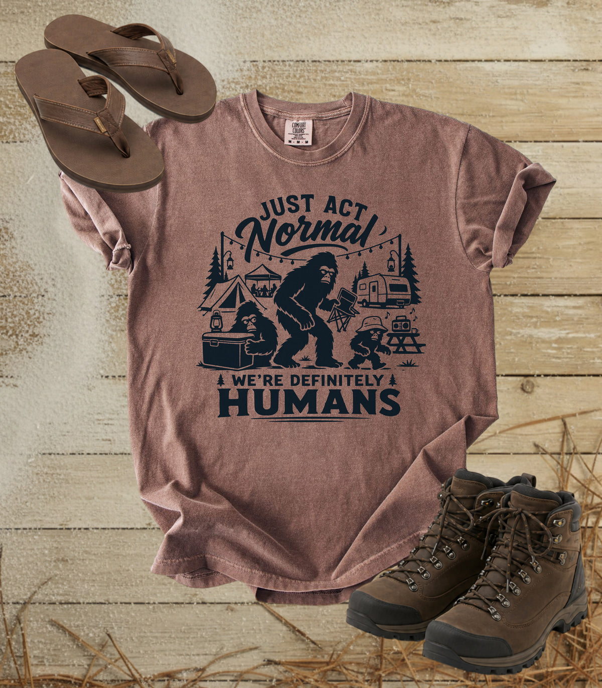 A comfort colors espresso color graphic t-shirt featuring a dark print of a Bigfoot family camping scene with the text 'JUST ACT Normal WE'RE DEFINITELY HUMANS'. The shirt is styled in a flat-lay arrangement on a rustic wooden background, accompanied by brown leather sandals and hiking boots.