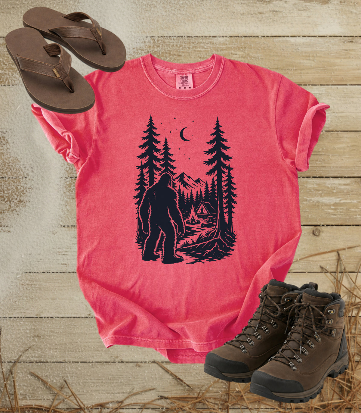 A comfort colors watermelon color short-sleeve t-shirt with a dark graphic of a Bigfoot in a pine forest at night looking at your camp, styled in a flat lay with brown flip-flops and hiking boots on a rustic wood background.