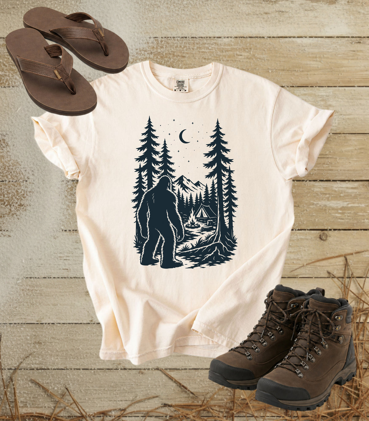A comfort colors ivory color short-sleeve t-shirt with a dark graphic of a Bigfoot in a pine forest at night looking at your camp, styled in a flat lay with brown flip-flops and hiking boots on a rustic wood background.