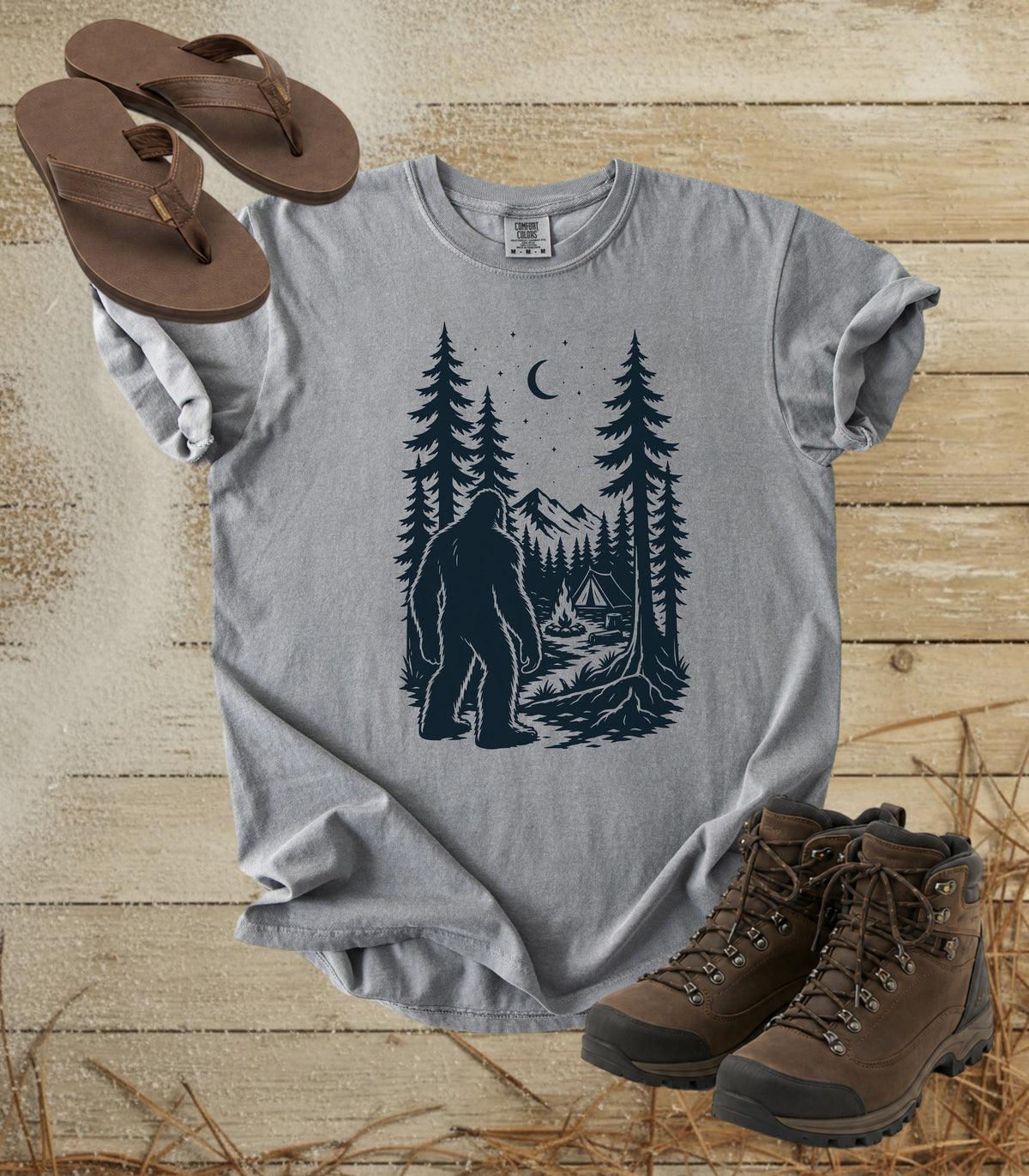A comfort colors grey color short-sleeve t-shirt with a dark graphic of a Bigfoot in a pine forest at night looking at your camp, styled in a flat lay with brown flip-flops and hiking boots on a rustic wood background.