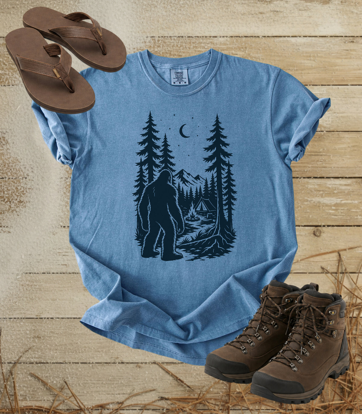 A comfort colors bluejean color short-sleeve t-shirt with a dark graphic of a Bigfoot in a pine forest at night looking at your camp, styled in a flat lay with brown flip-flops and hiking boots on a rustic wood background.