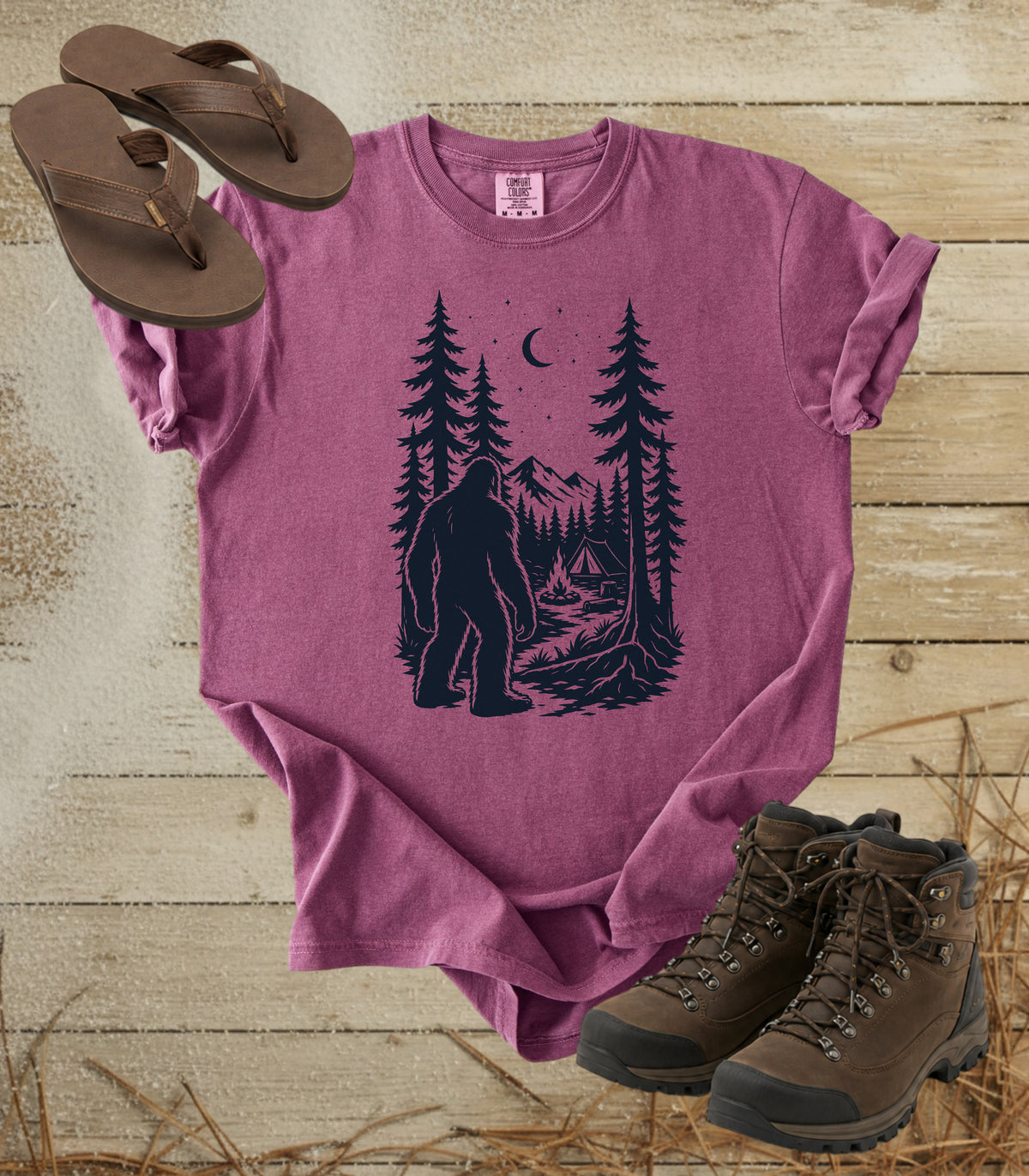 A comfort colors berry color short-sleeve t-shirt with a dark graphic of a Bigfoot in a pine forest at night looking at your camp, styled in a flat lay with brown flip-flops and hiking boots on a rustic wood background.