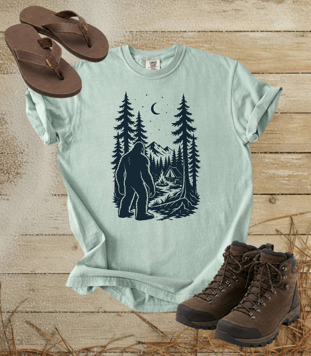 A comfort colors bay color short-sleeve t-shirt with a dark graphic of a Bigfoot in a pine forest at night looking at your camp, styled in a flat lay with brown flip-flops and hiking boots on a rustic wood background.