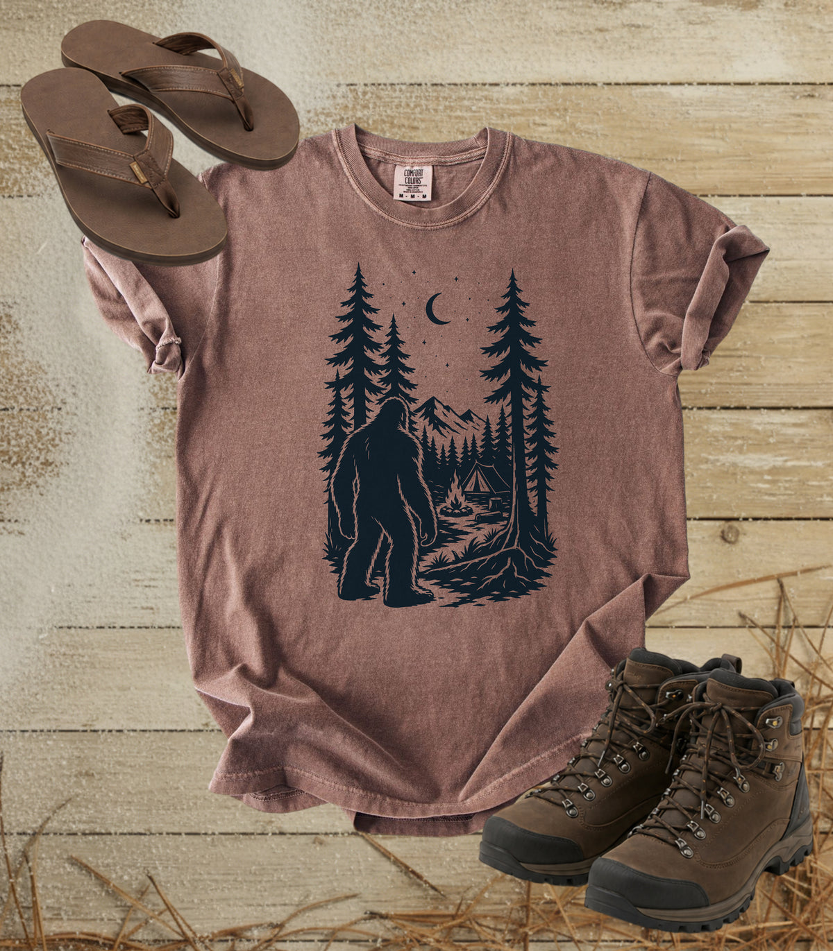 A comfort colors espresso color short-sleeve t-shirt with a dark graphic of a Bigfoot in a pine forest at night looking at your camp, styled in a flat lay with brown flip-flops and hiking boots on a rustic wood background.
