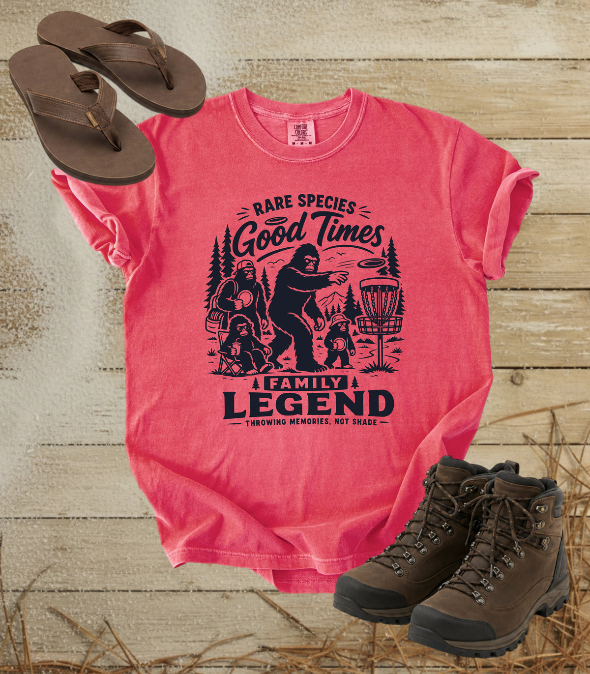 A comfort colors watermelon color graphic t-shirt featuring a dark blue print of Bigfoot and a family playing disc golf, with the text 'RARE SPECIES Good Times  FAMILY LEGEND, Throwing memories, not shade'. The shirt is styled in a flat-lay arrangement on a rustic wood background, accompanied by brown leather sandals and hiking boots.
