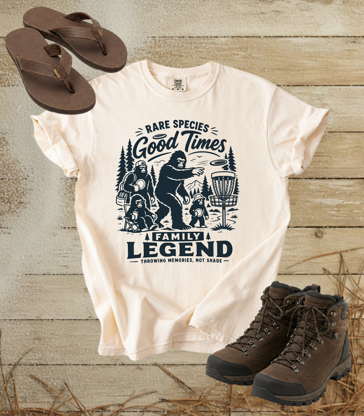 A comfort colors ivory color graphic t-shirt featuring a dark blue print of Bigfoot and a family playing disc golf, with the text 'RARE SPECIES Good Times  FAMILY LEGEND, Throwing memories, not shade'. The shirt is styled in a flat-lay arrangement on a rustic wood background, accompanied by brown leather sandals and hiking boots.