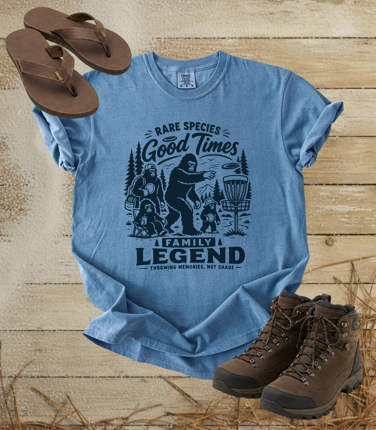 A comfort colors bluejean color graphic t-shirt featuring a dark blue print of Bigfoot and a family playing disc golf, with the text 'RARE SPECIES Good Times  FAMILY LEGEND, Throwing memories, not shade'. The shirt is styled in a flat-lay arrangement on a rustic wood background, accompanied by brown leather sandals and hiking boots.