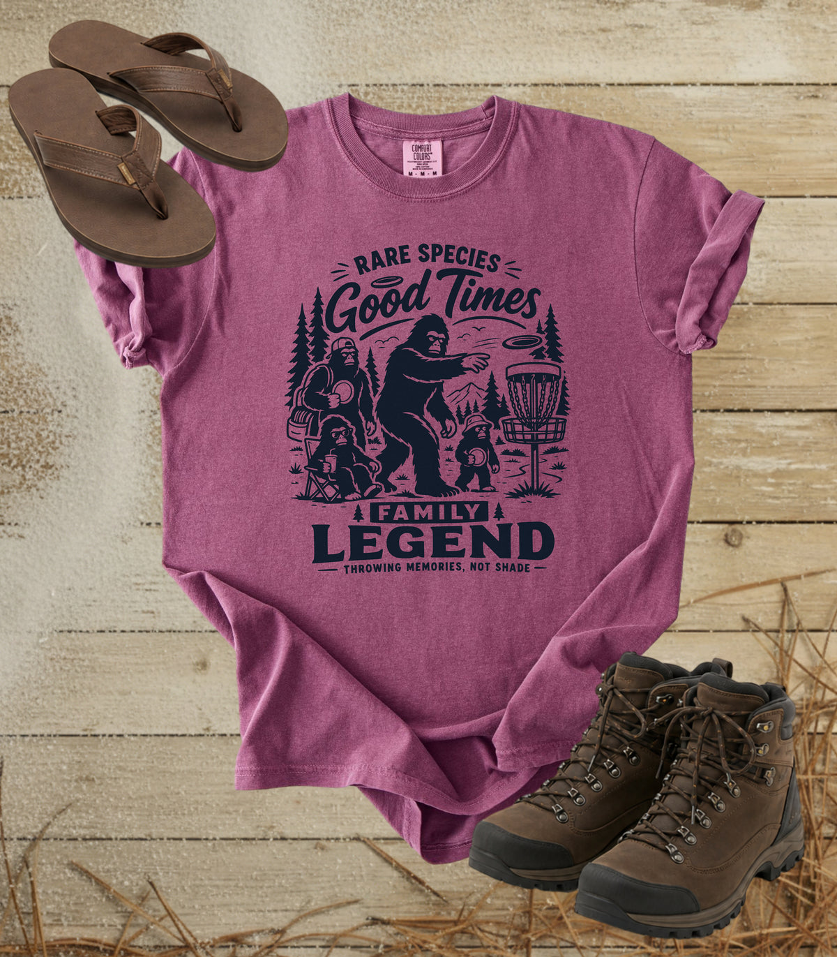 A comfort colors berry color graphic t-shirt featuring a dark blue print of Bigfoot and a family playing disc golf, with the text 'RARE SPECIES Good Times  FAMILY LEGEND, Throwing memories, not shade'. The shirt is styled in a flat-lay arrangement on a rustic wood background, accompanied by brown leather sandals and hiking boots.