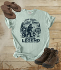 A comfort colors bay color graphic t-shirt featuring a dark blue print of Bigfoot and a family playing disc golf, with the text 'RARE SPECIES Good Times  FAMILY LEGEND, Throwing memories, not shade'. The shirt is styled in a flat-lay arrangement on a rustic wood background, accompanied by brown leather sandals and hiking boots.