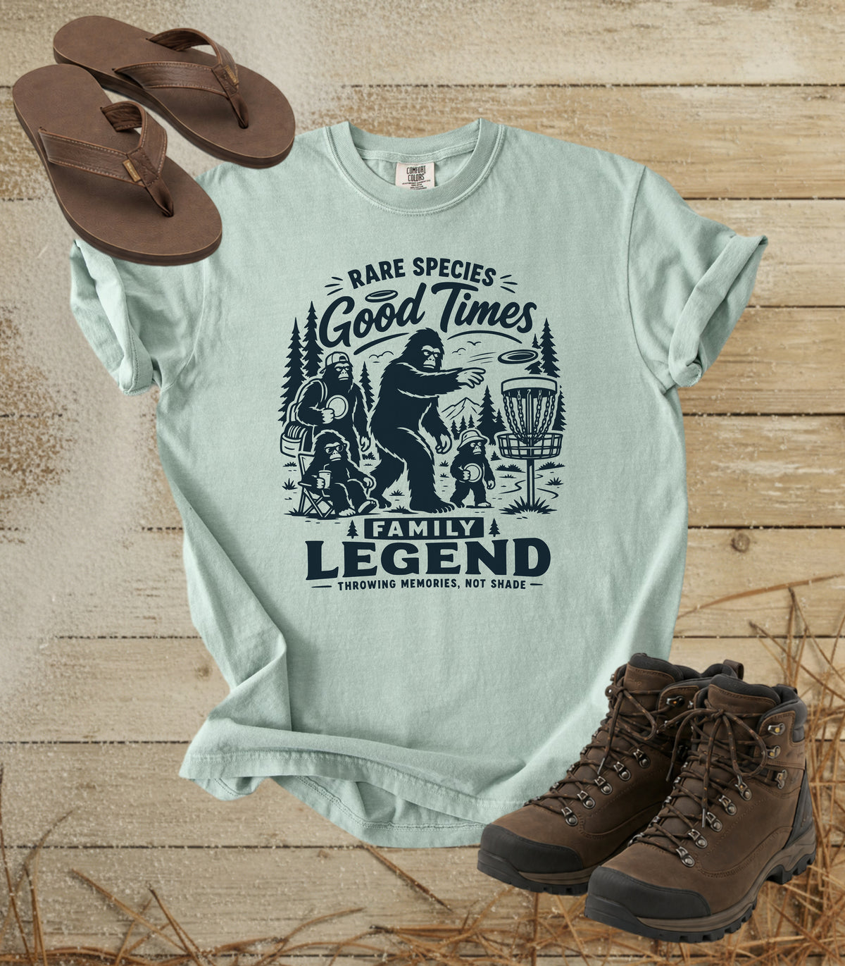 A comfort colors bay color graphic t-shirt featuring a dark blue print of Bigfoot and a family playing disc golf, with the text 'RARE SPECIES Good Times  FAMILY LEGEND, Throwing memories, not shade'. The shirt is styled in a flat-lay arrangement on a rustic wood background, accompanied by brown leather sandals and hiking boots.