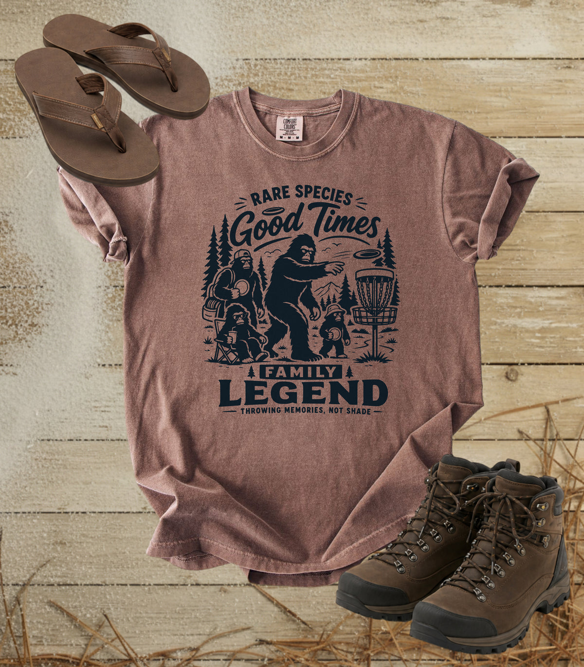A comfort colors espresso color graphic t-shirt featuring a dark blue print of Bigfoot and a family playing disc golf, with the text 'RARE SPECIES Good Times  FAMILY LEGEND, Throwing memories, not shade'. The shirt is styled in a flat-lay arrangement on a rustic wood background, accompanied by brown leather sandals and hiking boots.