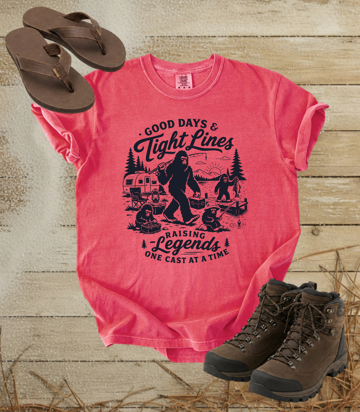 A comfort colors watermelon color graphic T-shirt laid flat on a rustic wood background, styled with brown flip-flops and hiking boots. The shirt features a dark blue illustrated graphic of a Bigfoot fishing scene with the text 'Good Days & Tight Lines' and 'Raising Legends One Cast at a Time.
