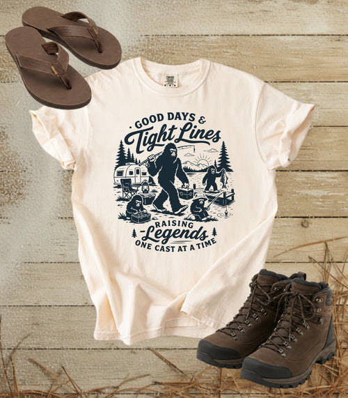A comfort colors ivory color graphic T-shirt laid flat on a rustic wood background, styled with brown flip-flops and hiking boots. The shirt features a dark blue illustrated graphic of a Bigfoot fishing scene with the text 'Good Days & Tight Lines' and 'Raising Legends One Cast at a Time.