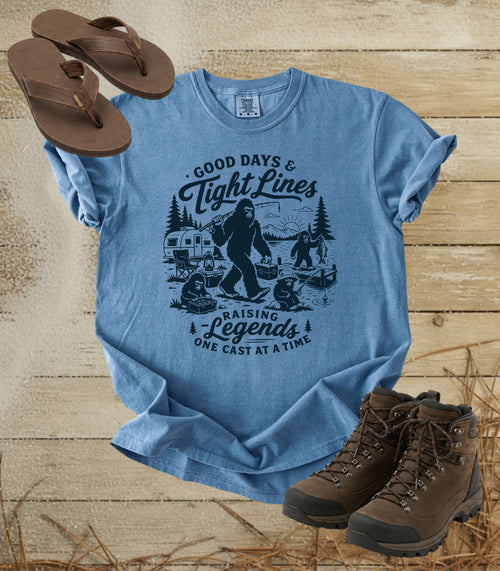 A comfort colors bluejean color graphic T-shirt laid flat on a rustic wood background, styled with brown flip-flops and hiking boots. The shirt features a dark blue illustrated graphic of a Bigfoot fishing scene with the text 'Good Days & Tight Lines' and 'Raising Legends One Cast at a Time.