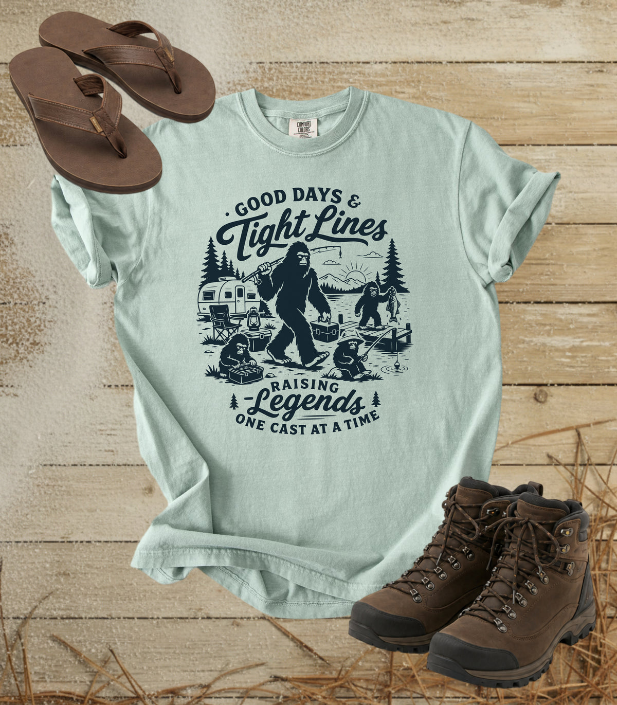 A comfort colors bay color graphic T-shirt laid flat on a rustic wood background, styled with brown flip-flops and hiking boots. The shirt features a dark blue illustrated graphic of a Bigfoot fishing scene with the text 'Good Days & Tight Lines' and 'Raising Legends One Cast at a Time.