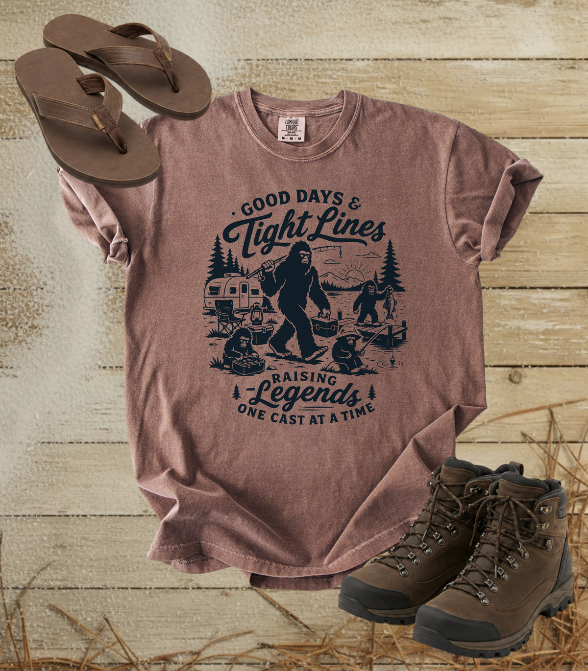 A comfort colors espresso color graphic T-shirt laid flat on a rustic wood background, styled with brown flip-flops and hiking boots. The shirt features a dark blue illustrated graphic of a Bigfoot fishing scene with the text 'Good Days & Tight Lines' and 'Raising Legends One Cast at a Time.