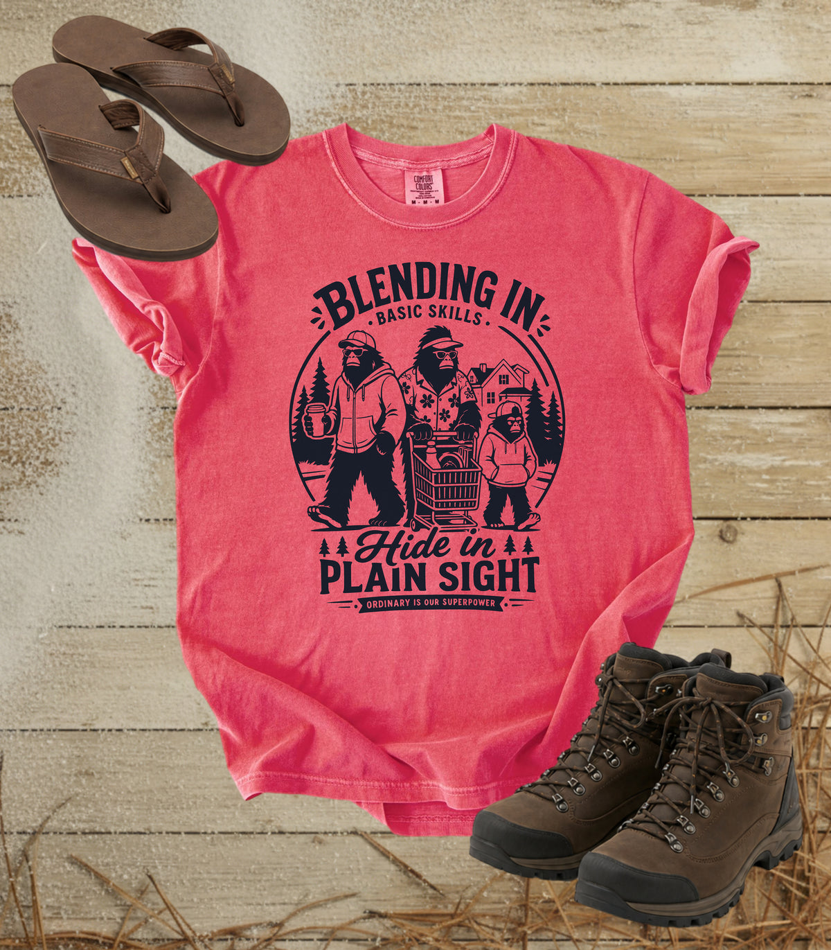 A comfort colors watermelon color graphic t-shirt featuring a dark blue bigfoot-themed outdoor print that reads 'Blending In: Basic Skills. Hide in Plain Sight.