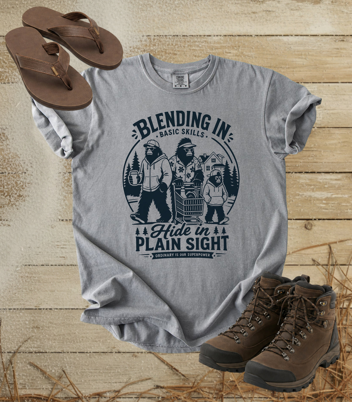 A comfort colors grey color graphic t-shirt featuring a dark blue bigfoot-themed outdoor print that reads 'Blending In: Basic Skills. Hide in Plain Sight.
