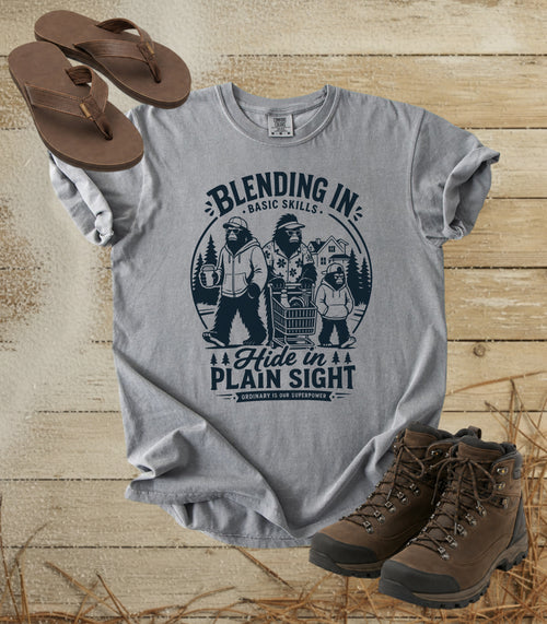 A comfort colors grey color graphic t-shirt featuring a dark blue bigfoot-themed outdoor print that reads 'Blending In: Basic Skills. Hide in Plain Sight.