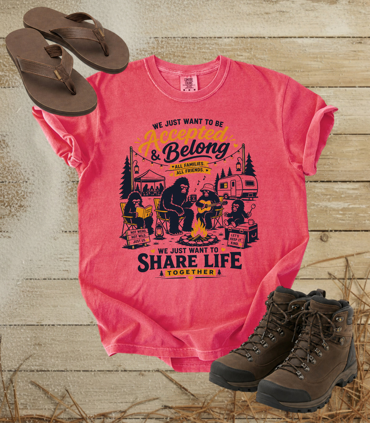 A comfort colors watermelon color graphic t-shirt laid flat on a rustic wood background, styled with brown flip-flops and hiking boots. The shirt features a black and yellow bigfoot camping scene with the text 'We Just Want to Be & Belong & Share Life Together'.
