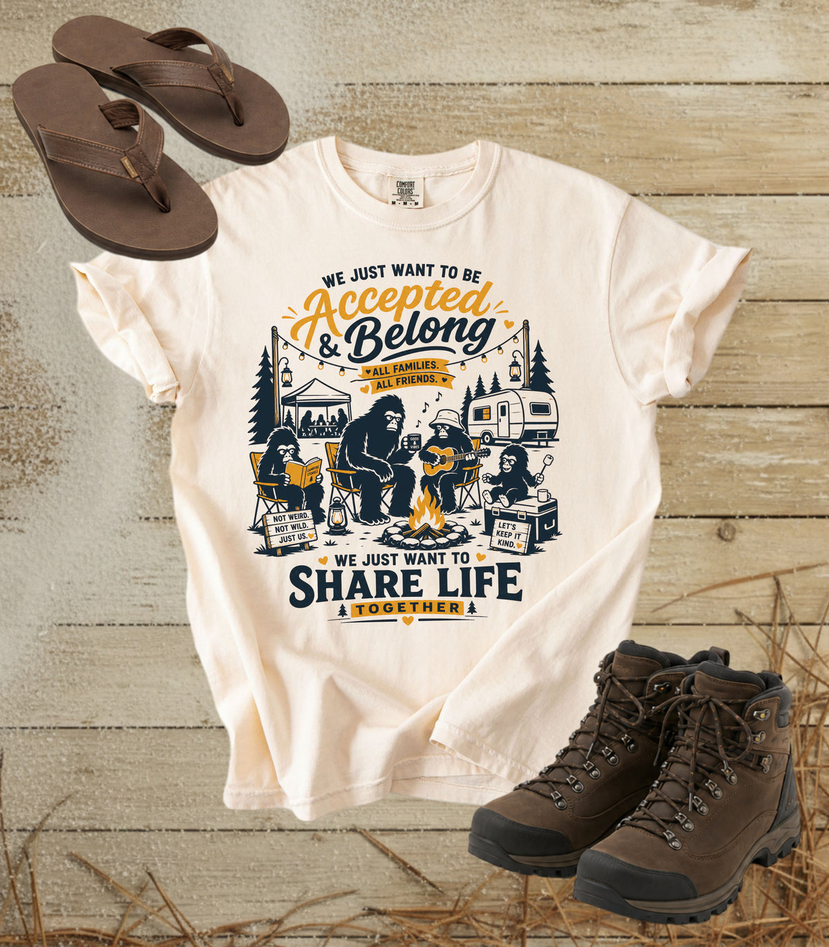 A comfort colors ivory color graphic t-shirt laid flat on a rustic wood background, styled with brown flip-flops and brown hiking boots. The t-shirt features a detailed camping scene with a bear and the text 'WE JUST WANT TO BE Accepted & Belong & SHARE LIFE & VEGAN THEM'.