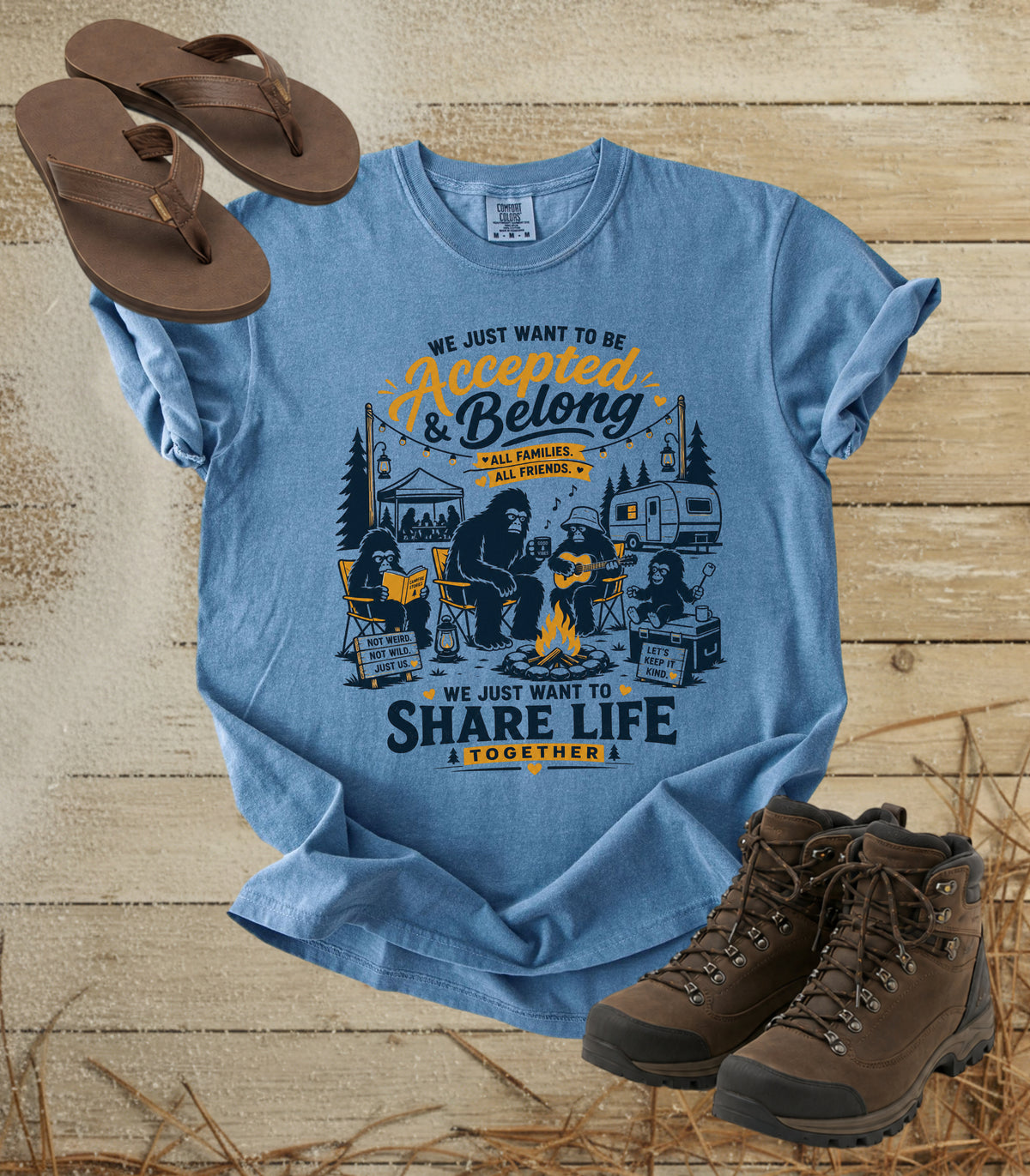 A comfort colors bluejean color graphic T-shirt laid flat on a rustic wood surface, styled with brown flip-flops and hiking boots. The shirt features a large front print with bigfoot camping imagery and the text 'We Just Want to Be Accepted & Belong - All Around My Family' and 'We Just Want to Share Life Together.