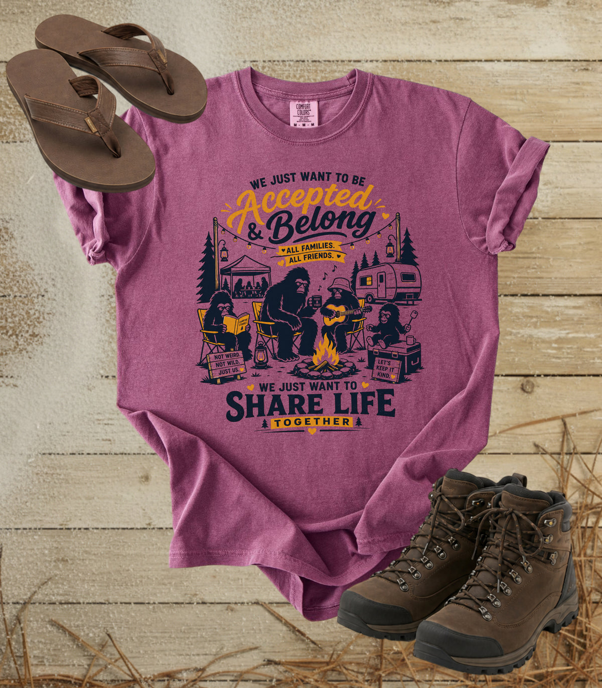 A comfort colors berry color graphic t-shirt laid flat on a rustic wood background, styled with brown flip-flops and hiking boots. The shirt features a camping-themed illustration of Bigfoot and other campers with the text 'WE JUST WANT TO BE Accepted & Belong ALL GEARLESS ALL TOGETHER WE JUST WANT TO SHARE LIFE TOGETHER'.