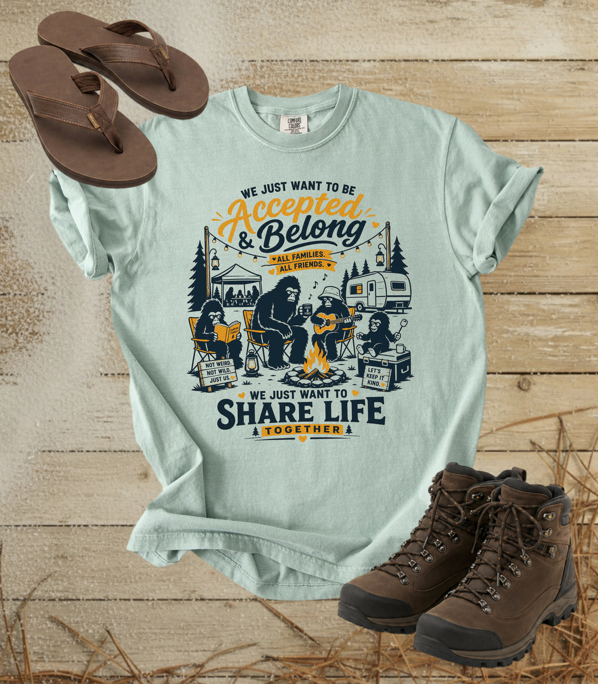 A comfort colors bay color graphic t-shirt laid flat on a rustic wood background, styled with brown flip-flops and hiking boots. The t-shirt features a large camping-themed graphic with a bear, tent, and campfire, accompanied by the text 'WE JUST WANT TO BE Accepted & Belong & SHARE LIFE TOGETHER!'.