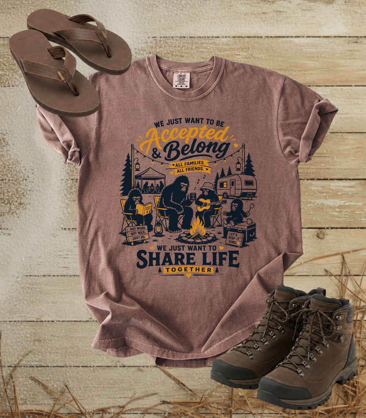 A comfort colors espresso color graphic t-shirt laid flat on a rustic wood background, styled with brown flip-flops and hiking boots. The shirt features a camping-themed graphic with the text 'WE JUST WANT TO BE Accepted & Belong & WE JUST WANT TO SHARE LIFE & TOGETHER A' in yellow and navy blue.