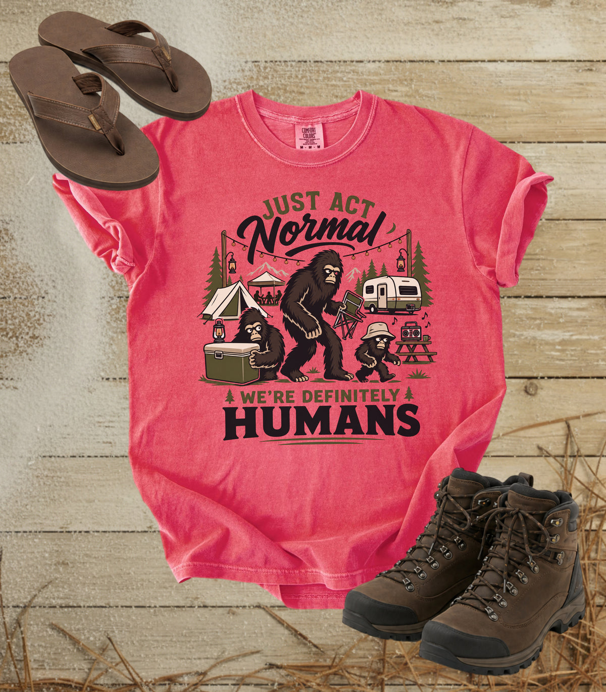 A comfort colors watermelon color graphic t-shirt laid flat on a rustic wood background, styled with brown flip-flops and hiking boots. The shirt features a camping-themed illustration of a Bigfoot and a woman with a dog, with the text 'JUST ACT Normal & WE'RE DEFINITELY HUMANS'.