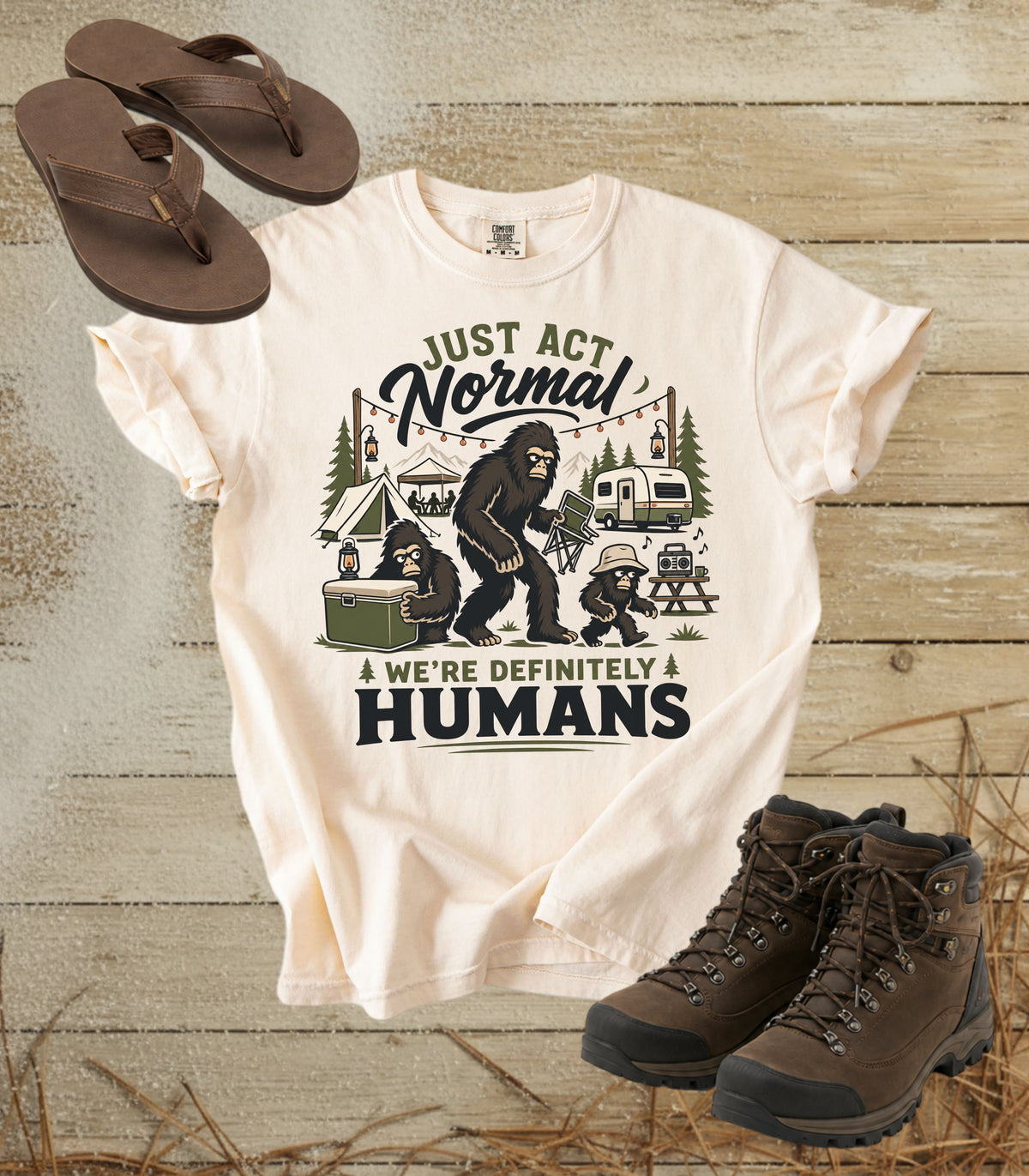 A comfort colors ivory color graphic t-shirt displayed in a flat-lay arrangement with brown flip-flops and hiking boots on a rustic wood background. The t-shirt features a humorous camping graphic with the text 'JUST ACT Normal A WE'RE DEFINITELY HUMANS' and an illustration of a Bigfoot family camping scene.