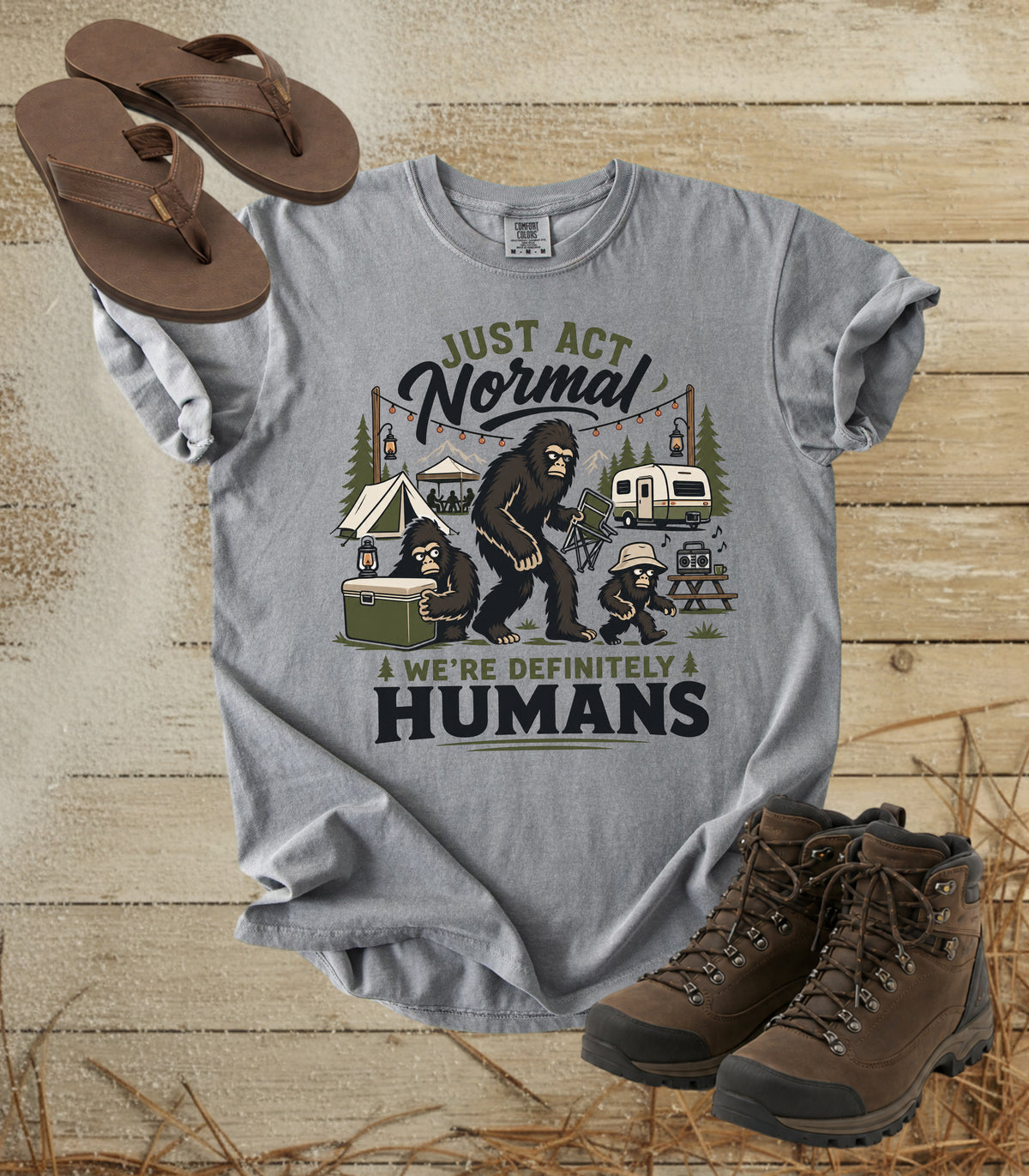 A comfort colors grey color graphic t-shirt featuring a Bigfoot camping scene with the text 'JUST ACT Normal WE'RE DEFINITELY HUMANS'. The shirt is styled in a flat-lay arrangement on a rustic wooden background alongside brown sandals and hiking boots.