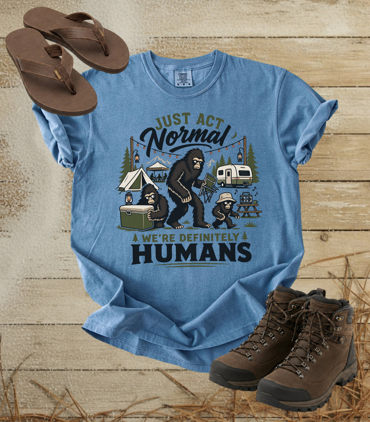 A comfort colors bluejean color graphic t-shirt laid flat on a rustic wood background, styled with brown flip-flops and hiking boots. The t-shirt features a cartoon graphic of a big cat-like figure and a child at a campsite with the text 'JUST ACT Normal WE'RE DEFINITELY HUMANS'.