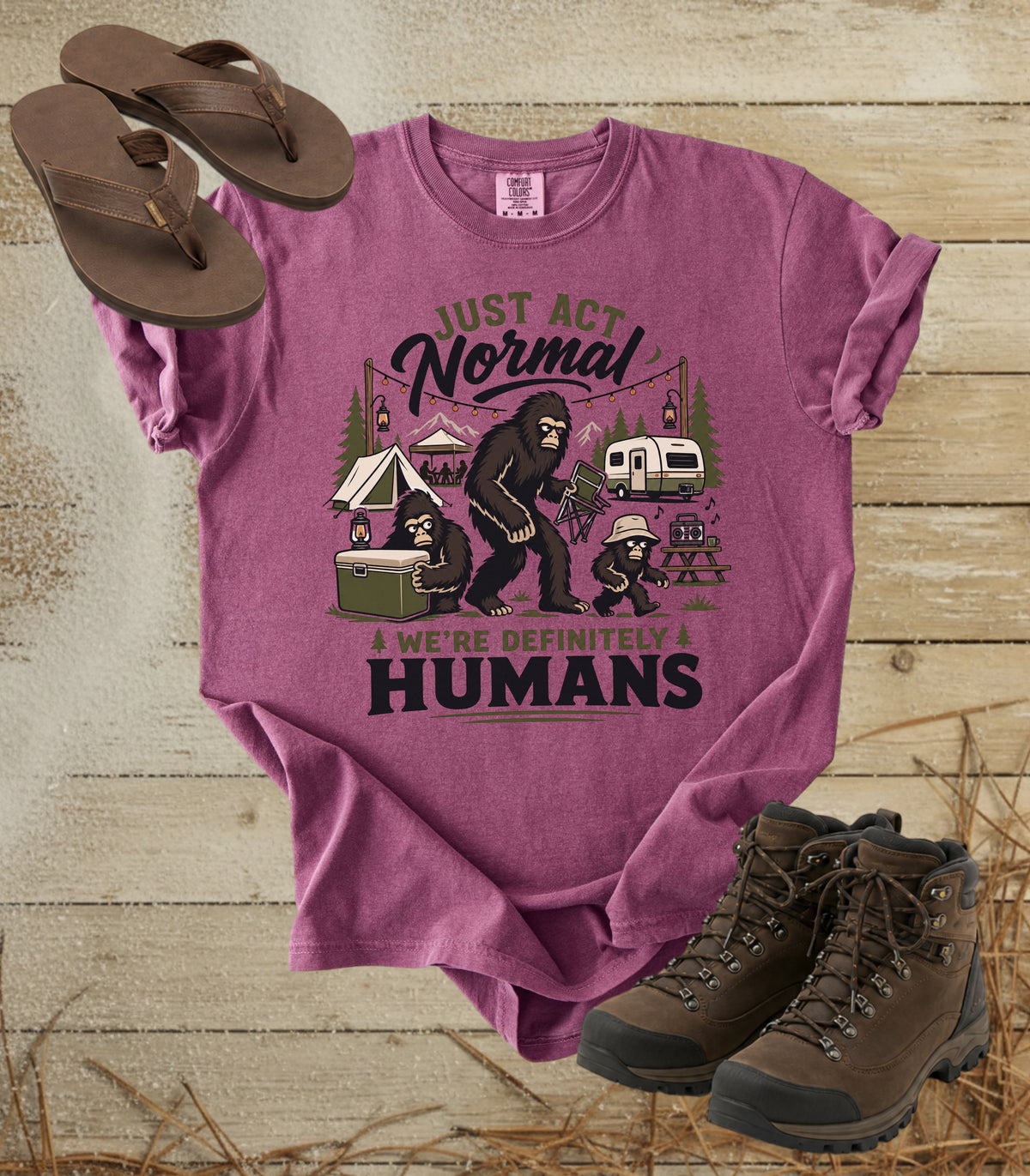 A comfort colors berry color graphic t-shirt featuring a Bigfoot camping scene with the text 'JUST ACT NORMAL' and 'WE'RE DEFINITELY HUMANS'. The shirt is styled in a flat lay on a rustic wood background, accompanied by brown flip-flops and hiking boots.