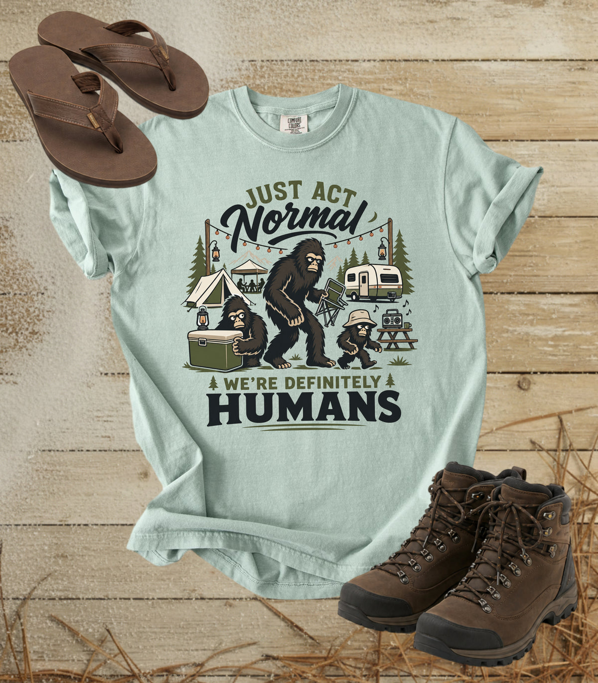 A comfort colors bay color graphic t-shirt displayed in a flat-lay arrangement with brown flip-flops and hiking boots on a rustic wood background. The t-shirt features a humorous camping graphic with a bigfoot, a child, a car, and the text 'JUST ACT Normal WE'RE DEFINITELY HUMANS'.