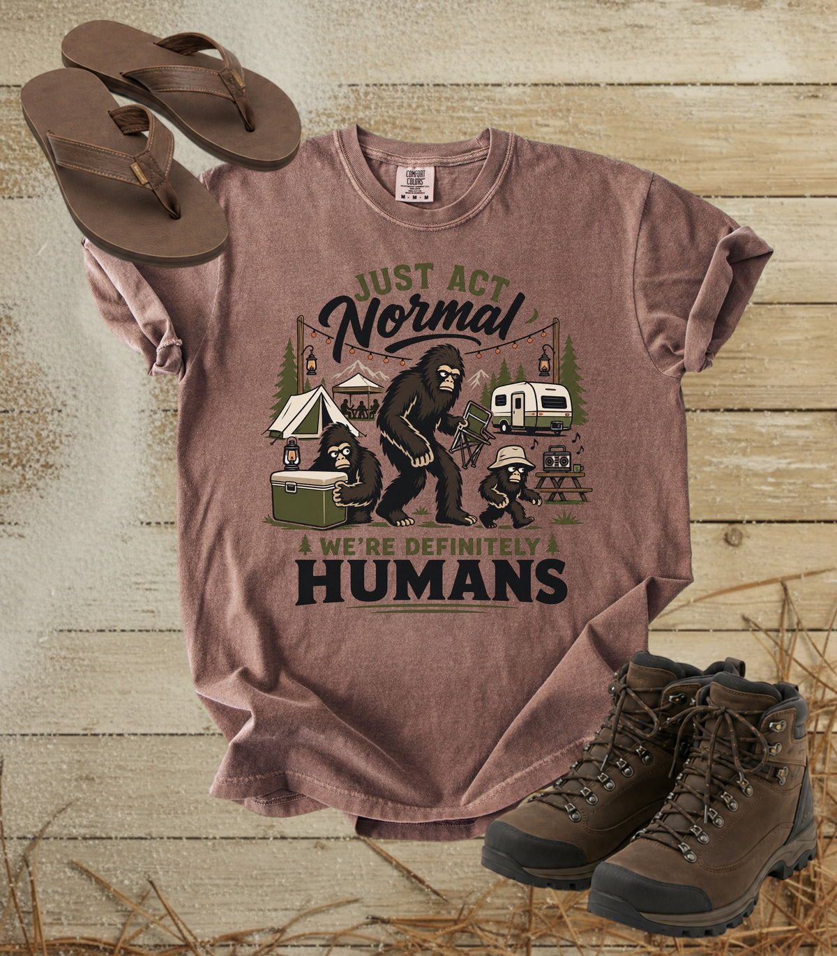 A comfort colors espresso color graphic t-shirt laid flat on a rustic wood background, styled with brown flip-flops and hiking boots. The shirt features a graphic of a bigfoot camping scene with the text 'JUST ACT Normal A WE'RE DEFINITELY HUMANS'.