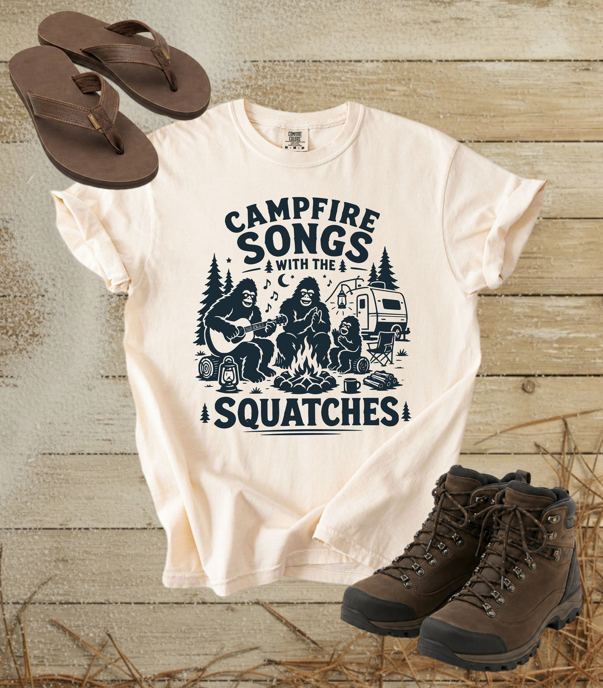 A comfort colors ivory color short-sleeve graphic t-shirt featuring a dark blue print of a campfire scene with the text 'CAMPFIRE SONGS WITH THE SQUATCHES'. The shirt is styled in a flat-lay arrangement on a rustic wood background, accompanied by brown flip-flops and hiking boots.