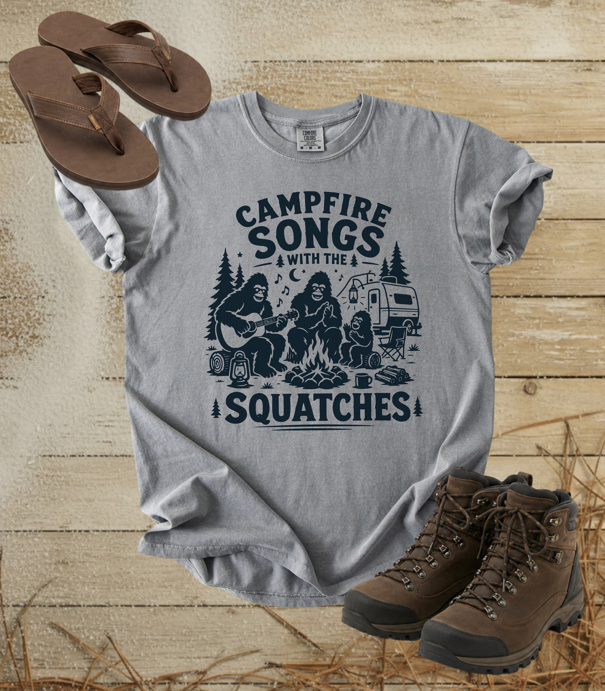 A comfort colors grey color graphic t-shirt laid flat on a rustic wood background, styled with brown flip-flops and hiking boots. The t-shirt features a dark navy blue graphic of a campfire scene with the text 'CAMPFIRE SONGS WITH THE SQUATCHES'.