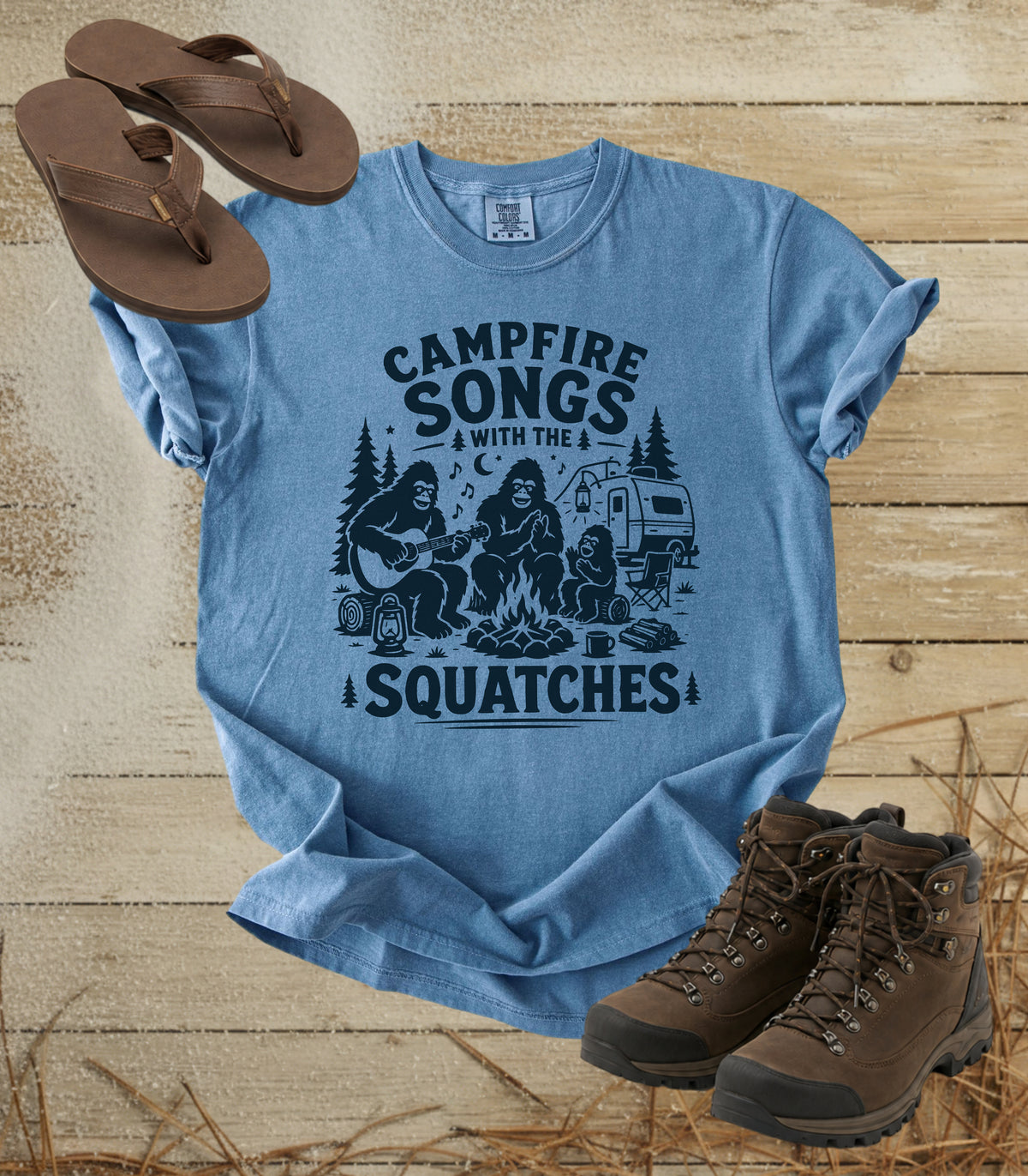 A comfort colors bluejean color graphic t-shirt featuring a black campfire design with the text 'CAMPFIRE SONGS WITH THE SQUATCHES'. The shirt is styled in a flat-lay arrangement on a rustic wooden background, accompanied by brown leather sandals, hiking boots, and straw.