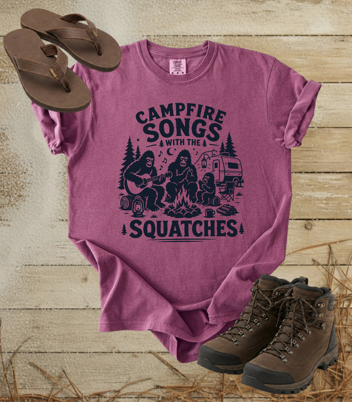 A comfort colors berry color t-shirt with a dark navy graphic print reading 'CAMPFIRE SONGS WITH THE SQUATCHES' and showing Bigfoot, a camper, and a campfire scene. The shirt is styled in a flat-lay on a rustic wood background with brown flip-flops and hiking boots.