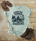A comfort colors bay color graphic t-shirt featuring a dark blue print of Bigfoot and a campfire scene with the text 'CAMPFIRE SONGS WITH THE SQUATCHES'. The shirt is styled in a flat-lay arrangement on a rustic wooden background, accompanied by brown leather sandals and hiking boots.
