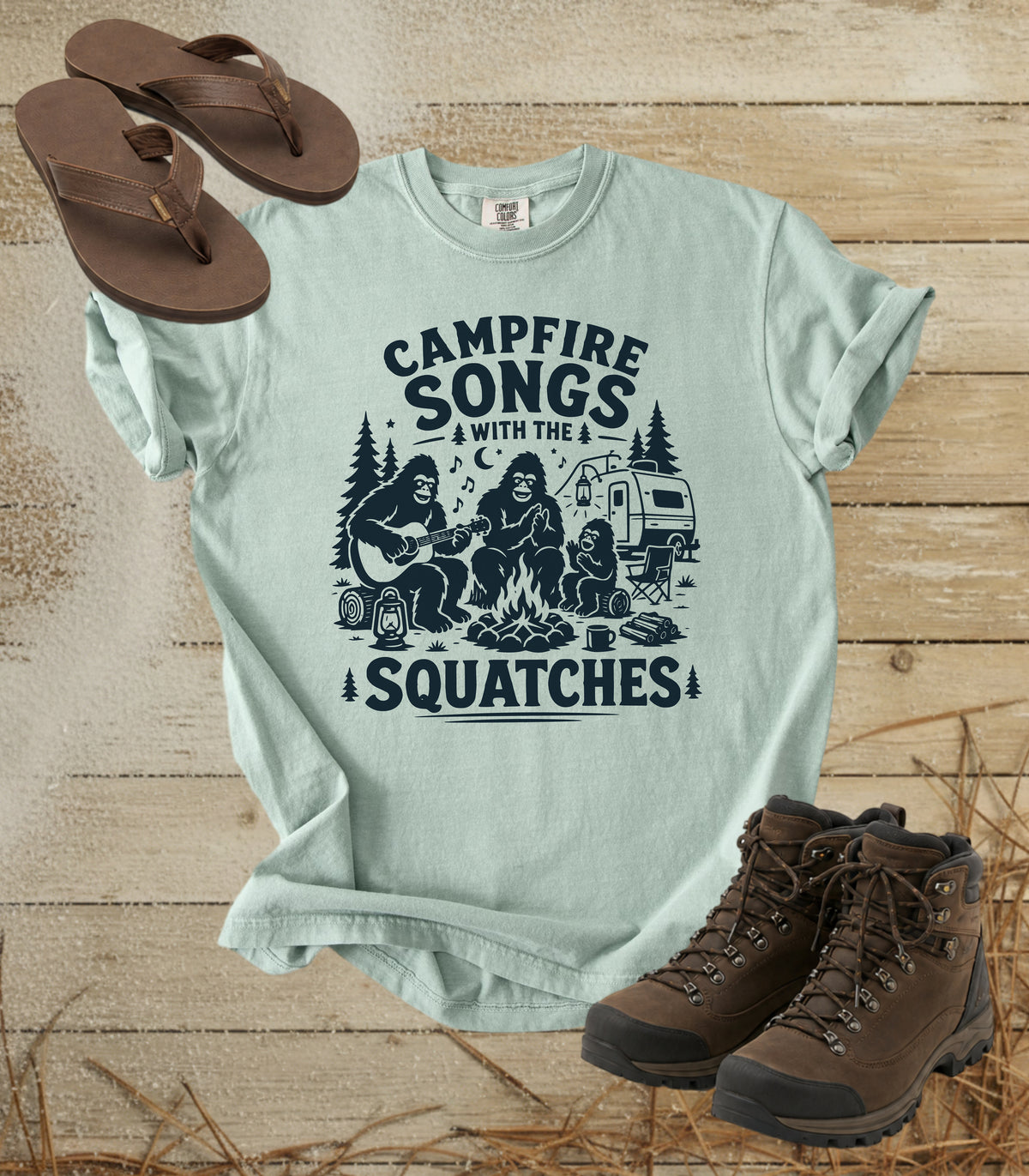 A comfort colors bay color graphic t-shirt featuring a dark blue print of Bigfoot and a campfire scene with the text 'CAMPFIRE SONGS WITH THE SQUATCHES'. The shirt is styled in a flat-lay arrangement on a rustic wooden background, accompanied by brown leather sandals and hiking boots.
