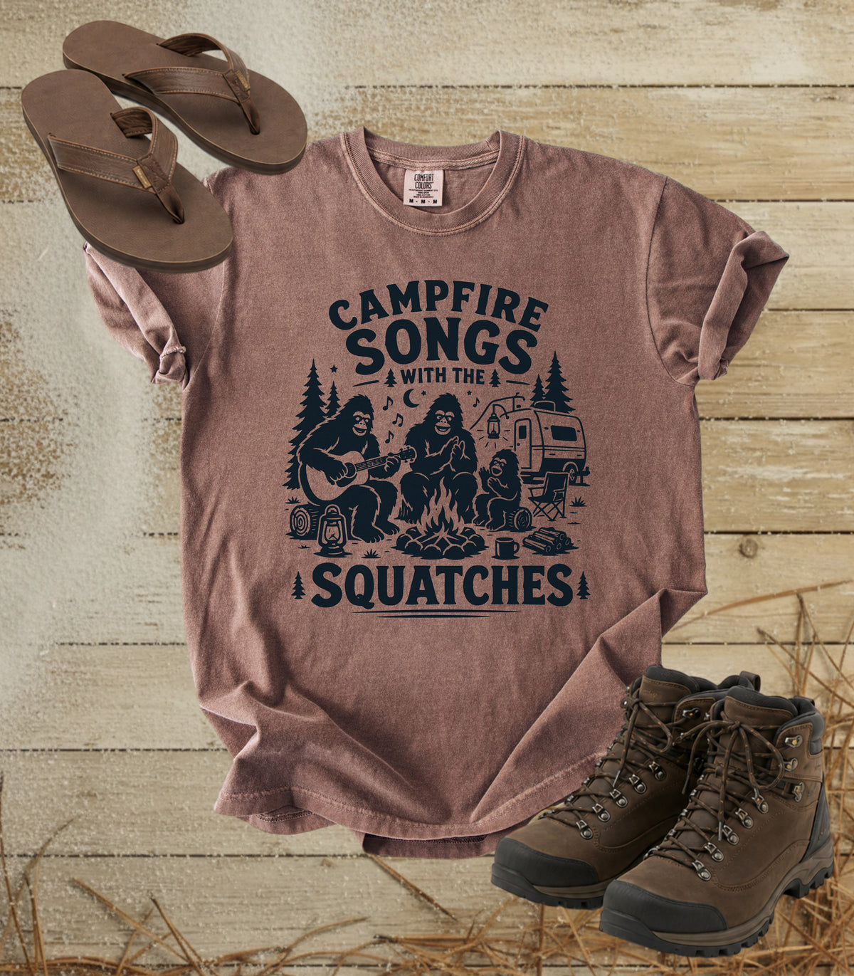 A comfort colors espresso color t-shirt with a dark blue graphic reading 'CAMPFIRE SONGS WITH THE SQUATCHES' and showing a campfire scene, styled in a flat lay with brown sandals, hiking boots, and straw on a rustic wood plank background.