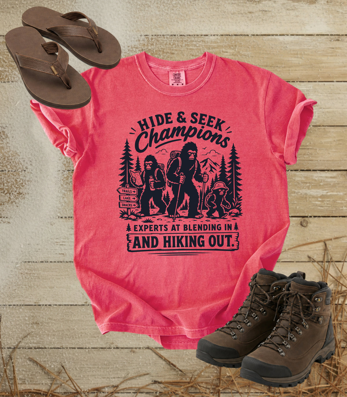 A comfort colors watermelon color short-sleeve graphic T-shirt laid flat on a rustic wood background, styled with brown flip-flops and hiking boots. The shirt has a black printed graphic and text reading 'Hide & Seek Champions' and 'Experts at blending in and hiking out.