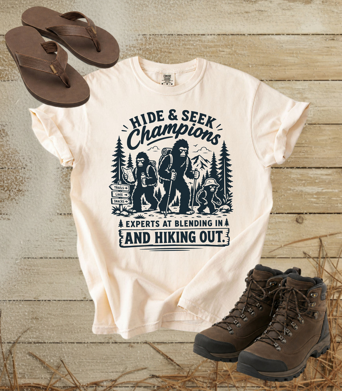 A comfort colors ivory color graphic t-shirt featuring a black print of Bigfoot and hiking scenes with the text 'HIDE & SEEK Champions & EXPERTS AT BLENDING IN AND HIKING OUT.' The shirt is styled in a flat-lay arrangement on a rustic wood background, accompanied by brown flip-flops and hiking boots.