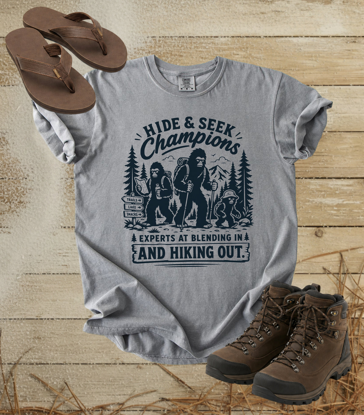 A comfort colors grey color graphic t-shirt featuring a dark blue 'Hide & Seek Champions' Bigfoot design, styled in a flat lay with brown sandals, hiking boots, and straw on a rustic wooden background.